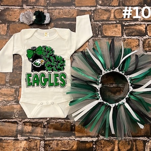 Könnte beinhalten: Ein cremefarbener Baby-Body mit dem Wort "EAGLES" in Grün und Schwarz, zusammen mit einem passenden Tutu-Rock und Stirnband. Der Body hat eine grün-schwarze Grafik. Das Tutu ist grün, schwarz und weiß.