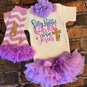 以下が含まれることがあります： ゴールドのグリッター文字で「Silly Rabbit Easter is for Jesus」と書かれた白いボディスーツ、パープルの花のヘッドバンド、ゴールドのグリッターのウサギが付いた白とパープルのストライプのレッグウォーマー、パープルのサテンのリボンが付いたパープルのチュールスカートを組み合わせた、女の子の赤ちゃん用イースターの衣装です。
