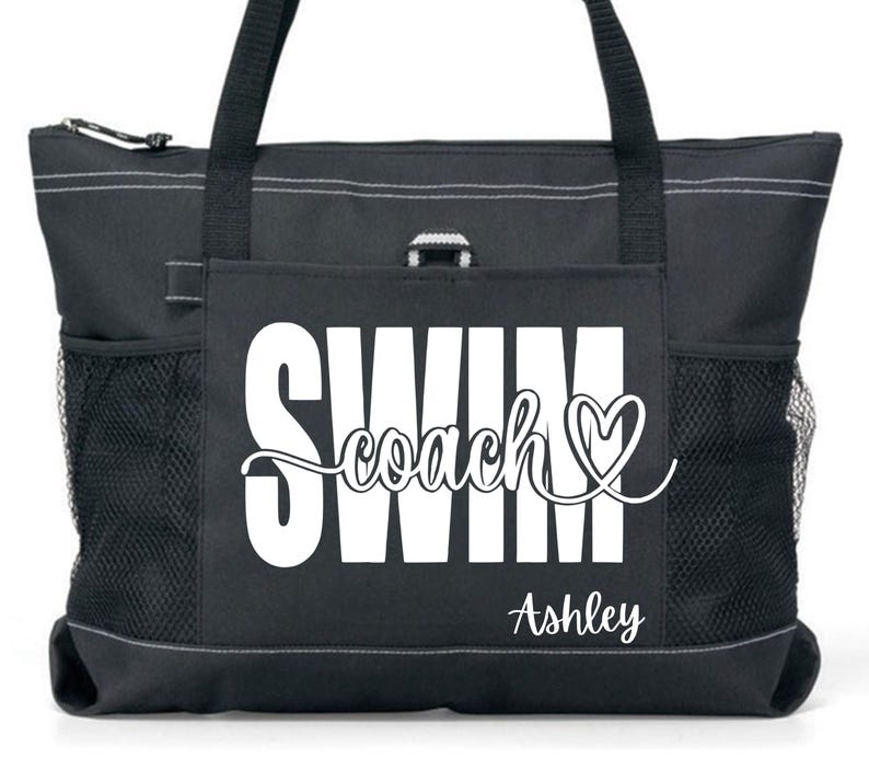 May include: Black tote bag with white text that reads "SWIM coach" with a heart symbol and the name "Ashley" below. The bag has two side mesh pockets, a zippered top, and black handles.