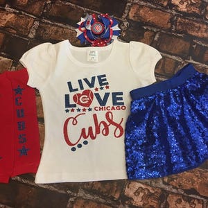 Peut inclure: Un t-shirt blanc avec le texte "Live Love Chicago Cubs" en paillettes rouges, bleues et blanches. Le t-shirt est associ&eacute; &agrave; une jupe paillet&eacute;e bleue et &agrave; des jambi&egrave;res ray&eacute;es rouges et blanches. Un n&oelig;ud bleu et blanc est plac&eacute; sur le haut du t-shirt.