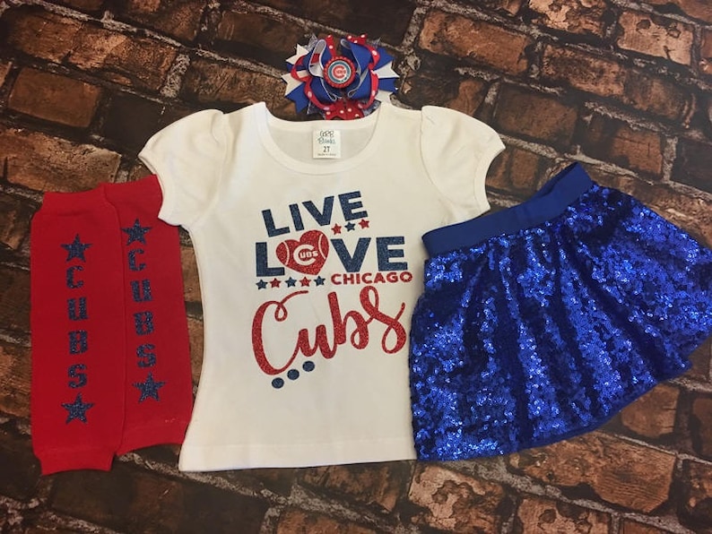 Peut inclure: Un t-shirt blanc avec le texte "Live Love Chicago Cubs" en paillettes rouges, bleues et blanches. Le t-shirt est associ&eacute; &agrave; une jupe paillet&eacute;e bleue et &agrave; des jambi&egrave;res ray&eacute;es rouges et blanches. Un n&oelig;ud bleu et blanc est plac&eacute; sur le haut du t-shirt.