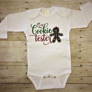 May include: White long-sleeve baby onesie with green and red glitter text that reads "The Cookie Tester" and a black gingerbread person graphic.