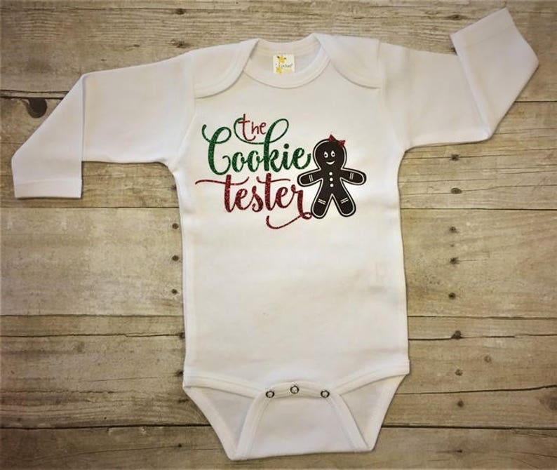 May include: White long-sleeve baby onesie with green and red glitter text that reads "The Cookie Tester" and a black gingerbread person graphic.