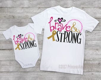 May include: Two white t-shirts, one a baby bodysuit, the other a standard tee. Both feature the words "brave & strong" in pink, black, and gold glitter, with a heart and ribbon design. The bodysuit is short-sleeved.