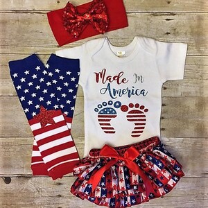 May include: A baby outfit set in red, white, and blue with a red headband, a white bodysuit with the text "Made in America" and a pair of red, white, and blue footprints, red, white, and blue leg warmers, and a red, white, and blue ruffled skirt with a red bow.