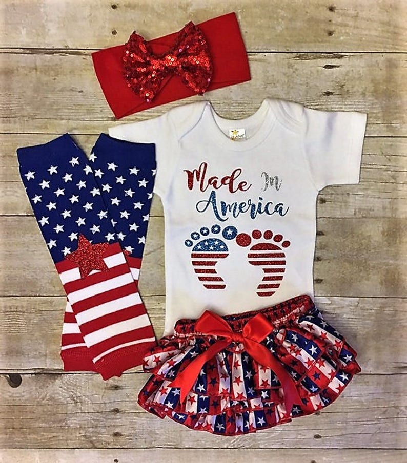 May include: A baby outfit set in red, white, and blue with a red headband, a white bodysuit with the text "Made in America" and a pair of red, white, and blue footprints, red, white, and blue leg warmers, and a red, white, and blue ruffled skirt with a red bow.