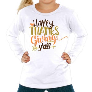 Puede incluir: Camiseta blanca de manga larga con el texto "Happy Thanks Giving y'all" en letras marrones, naranjas y doradas con un pequeño corazón y un gráfico de flecha.