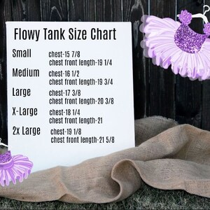 Puede incluir: Una tarjeta blanca con una tabla de tallas para tanques fluidos. La tabla enumera las tallas desde la peque&ntilde;a hasta la 2x grande con las medidas de pecho y longitud del pecho frontal correspondientes en pulgadas. La tarjeta est&aacute; sobre una superficie de arpillera marr&oacute;n.