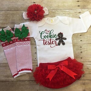 May include: A white baby onesie with the text "The Cookie Tester" and a gingerbread person graphic. The onesie is paired with a red tulle tutu, a red headband with a red flower, and white and red striped leg warmers with green bows.