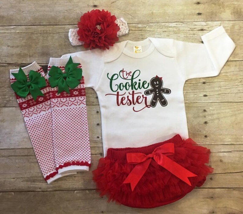 May include: A white baby onesie with the text "The Cookie Tester" and a gingerbread person graphic. The onesie is paired with a red tulle tutu, a red headband with a red flower, and white and red striped leg warmers with green bows.
