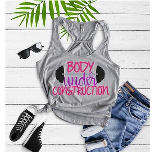Puede incluir: Camiseta de tirantes gris con el texto "BODY under CONSTRUCTION" en letras rosa y morado con un gr&aacute;fico de mancuerna negro sobre el texto.