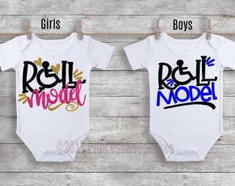 Może przedstawiać: Dwa białe body dla niemowląt z krótkimi rękawami. Body mają nadrukowany napis "Roll Model". Słowo "Roll" zawiera symbol wózka inwalidzkiego. Jedno body ma różowe cekiny, a drugie niebieskie.