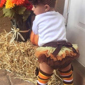Pu&ograve; includere: Una bambina che indossa una maglietta bianca, un tut&ugrave; di tulle marrone e giallo e scaldagambe a righe marroni e arancioni. Indossa una fascia per capelli con un fiore marrone e si trova vicino a una zucca e un mazzo di fiori arancioni e gialli.
