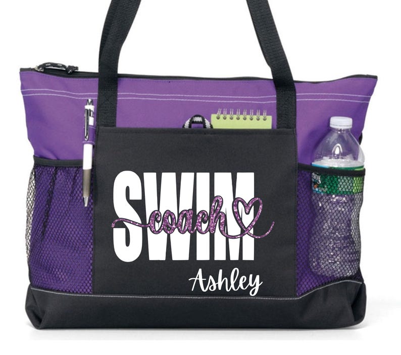 May include: A purple and black tote bag with the word "SWIM" in white block letters and "coach" in a glittery purple script. The bag has side pockets, a water bottle, and a pen. The name "Ashley" is printed below.