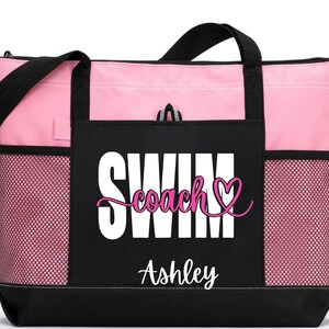 May include: A pink and black tote bag with the words "SWIM coach" and a heart graphic. The name "Ashley" is printed below the text. The bag has black handles and mesh side pockets.