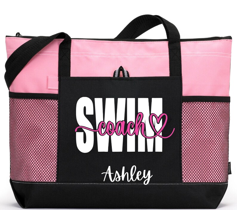 May include: A pink and black tote bag with the words "SWIM coach" and a heart graphic. The name "Ashley" is printed below the text. The bag has black handles and mesh side pockets.
