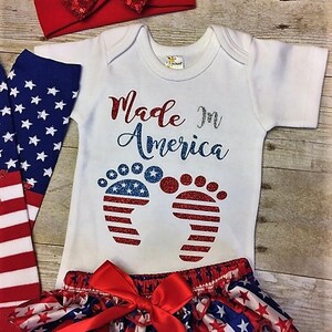 May include: A baby outfit set with a white bodysuit, red and blue star print ruffle bloomers, red and white headband, and blue and white star print socks. The bodysuit has the text "Made in America" with glitter accents and a pair of red, white, and blue glitter footprints.