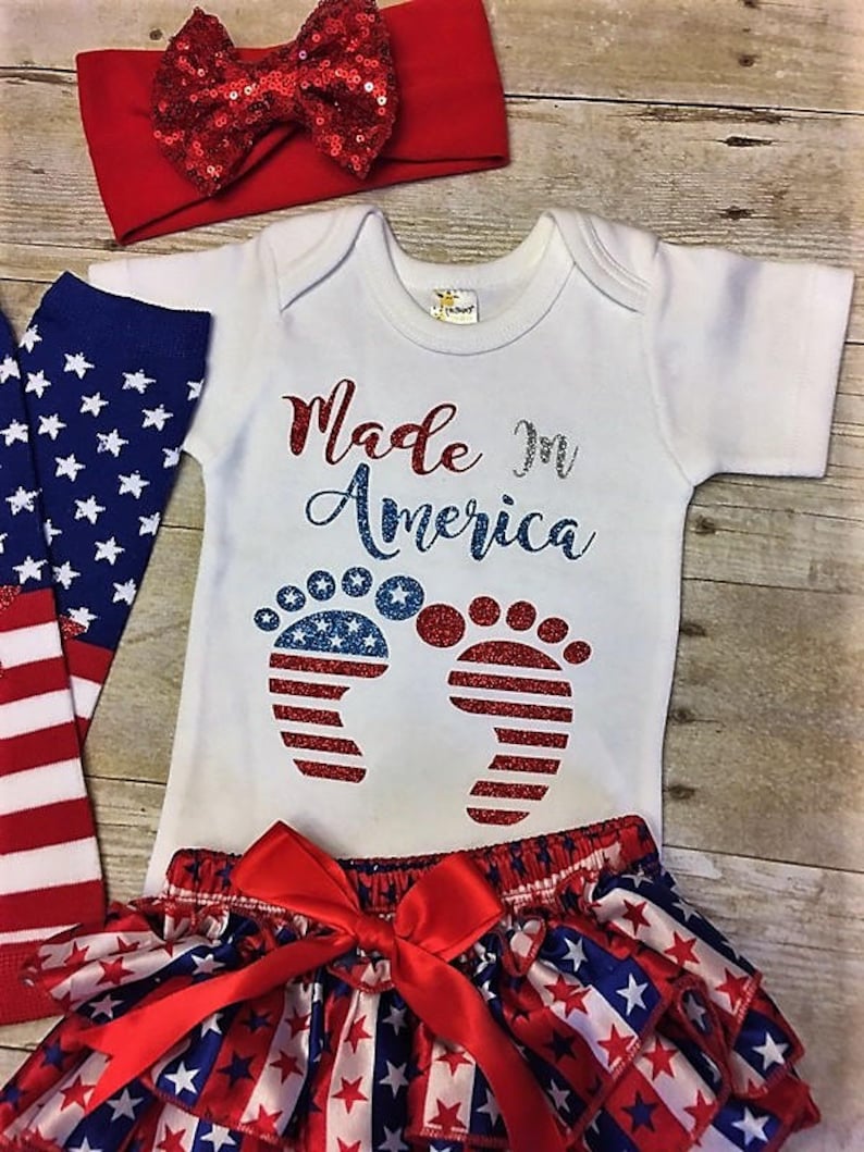 May include: A baby outfit set with a white bodysuit, red and blue star print ruffle bloomers, red and white headband, and blue and white star print socks. The bodysuit has the text "Made in America" with glitter accents and a pair of red, white, and blue glitter footprints.