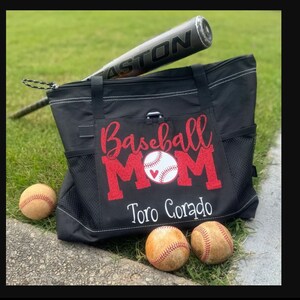 May include: Black tote bag with a red glitter design that says "Baseball Mom" with a white baseball and red heart. The bag has a white embroidered name "Toro Corado" and is sitting on a grassy surface with three baseballs.