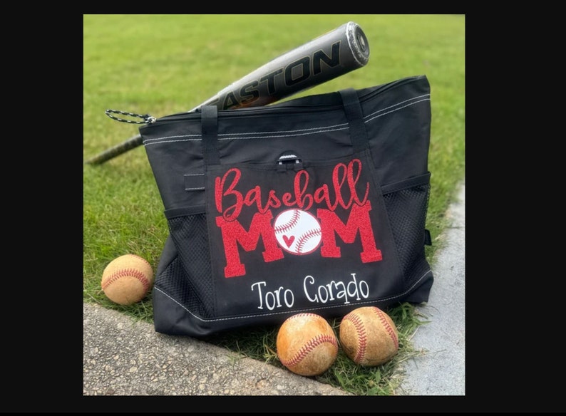 May include: Black tote bag with a red glitter design that says "Baseball Mom" with a white baseball and red heart. The bag has a white embroidered name "Toro Corado" and is sitting on a grassy surface with three baseballs.