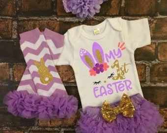 May include: A baby girl's Easter outfit with a white bodysuit that says "My 1st Easter" with bunny ears, a purple tulle skirt with a gold bow, purple and white chevron leg warmers with a gold bunny, and a purple flower headband.