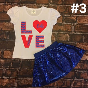 Peut inclure: Un t-shirt blanc avec un graphique rouge "LOVE" comportant un motif chevron bleu et un c&oelig;ur rouge avec le mot "Cubs" &agrave; l'int&eacute;rieur. Le t-shirt est associ&eacute; &agrave; une jupe paillet&eacute;e bleue.