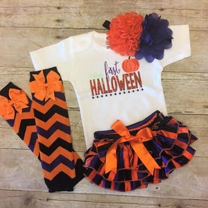 May include: A baby outfit for Halloween, featuring a white bodysuit with the text "first HALLOWEEN" and a pumpkin graphic, orange and purple striped bloomers with a bow, orange and purple chevron leg warmers with bows, and a headband with orange and purple flowers.