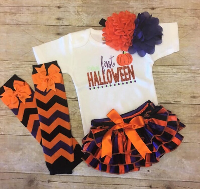 May include: A baby outfit for Halloween, featuring a white bodysuit with the text "first HALLOWEEN" and a pumpkin graphic, orange and purple striped bloomers with a bow, orange and purple chevron leg warmers with bows, and a headband with orange and purple flowers.