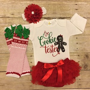 May include: A white onesie with the text "The Cookie Tester" and a gingerbread person graphic. The onesie is paired with a red tulle tutu, green and red leg warmers, and a red flower headband.