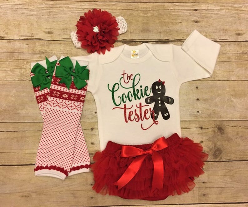 May include: A white onesie with the text "The Cookie Tester" and a gingerbread person graphic. The onesie is paired with a red tulle tutu, green and red leg warmers, and a red flower headband.