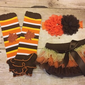 Pu&ograve; includere: Un scaldagambe a righe marroni e arancioni con un volant marrone e un tut&ugrave; di tulle marrone e arancione con un fiocco di raso marrone. Il tut&ugrave; ha una cintura marrone ed &egrave; decorato con un fiore marrone.