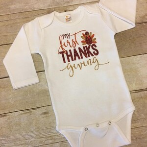 Pu&ograve; includere: Body bianco a maniche lunghe per neonato con il testo "my first THANKS giving" in glitter dorato e una grafica di tacchino in glitter rosso, arancione e dorato.