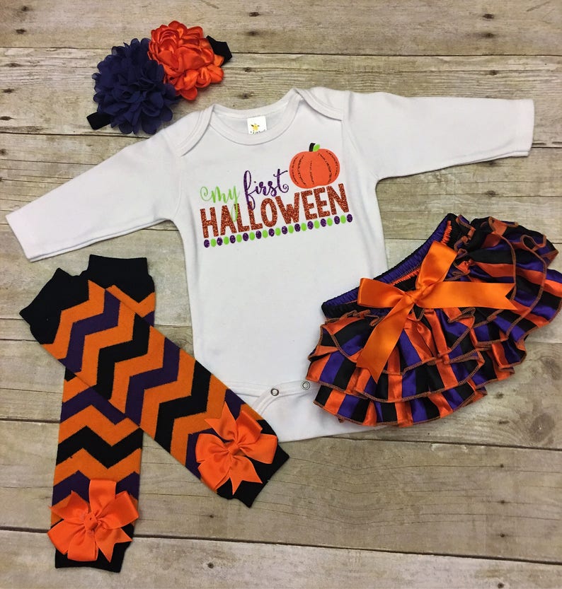 May include: A baby outfit set for Halloween, featuring a white long-sleeved bodysuit with the text "My first Halloween" and a pumpkin graphic, orange and black striped ruffle bloomers, and orange and black chevron leg warmers with orange bows.