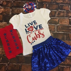 Peut inclure: Un t-shirt blanc avec le texte "Live Love Chicago Cubs" en paillettes rouges, bleues et blanches. Le t-shirt est associ&eacute; &agrave; une jupe en sequins bleus et &agrave; des jambi&egrave;res rouges et bleues avec des &eacute;toiles blanches et le mot "Cubs" imprim&eacute; dessus. Un n&oelig;ud rouge, blanc et bleu est &eacute;galement inclus.