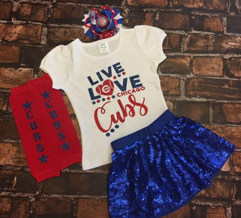 Peut inclure: Un t-shirt blanc avec le texte "Live Love Chicago Cubs" en paillettes rouges, bleues et blanches. Le t-shirt est associ&eacute; &agrave; une jupe en sequins bleus et &agrave; des jambi&egrave;res rouges et bleues avec des &eacute;toiles blanches et le mot "Cubs" imprim&eacute; dessus. Un n&oelig;ud rouge, blanc et bleu est &eacute;galement inclus.