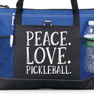 May include: A blue and black tote bag with a zippered top and mesh side pockets. The bag has a black pocket on the front with the text "Peace. Love. Pickleball." in white.
