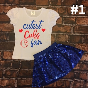 Peut inclure: T-shirt blanc avec inscription rouge et bleue "cutest Cubs fan" avec un ballon de baseball et deux c&oelig;urs rouges. Le t-shirt est associ&eacute; &agrave; une jupe bleue &agrave; paillettes.