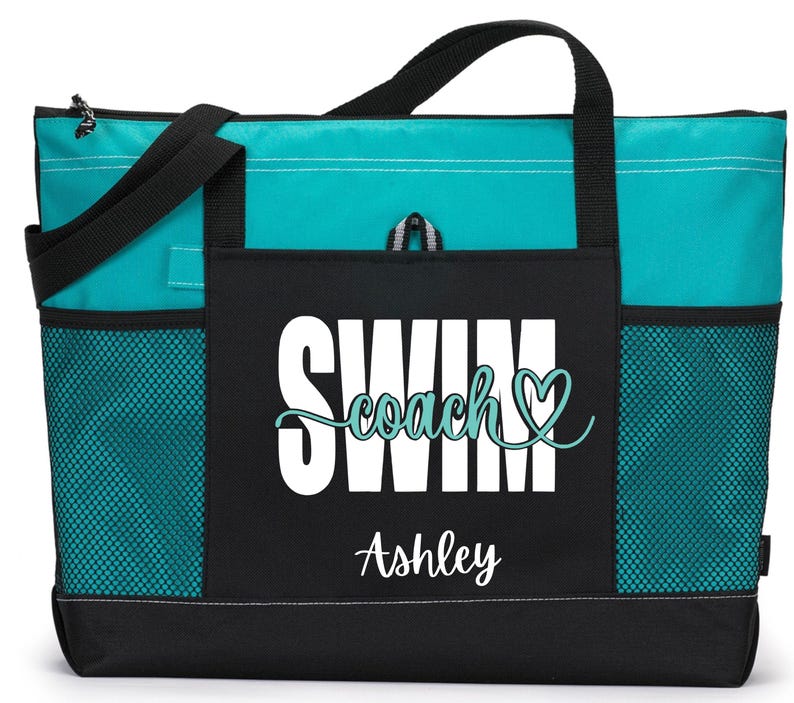 May include: A teal and black tote bag with the words "SWIM coach" and the name "Ashley" printed on the front. The bag has black handles, side pockets, and a zippered top. The bag is designed for carrying items.