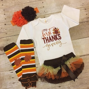 Pu&ograve; includere: Un set di abbigliamento per neonati per il Ringraziamento. Include un body bianco a maniche lunghe con il testo "my first THANKS giving" in glitter dorato, un gambaletto a righe marroni e arancioni con un volant marrone, una gonna di tulle marrone con strati di tulle arancioni e gialli, e una fascia per capelli con fiori marroni e arancioni.