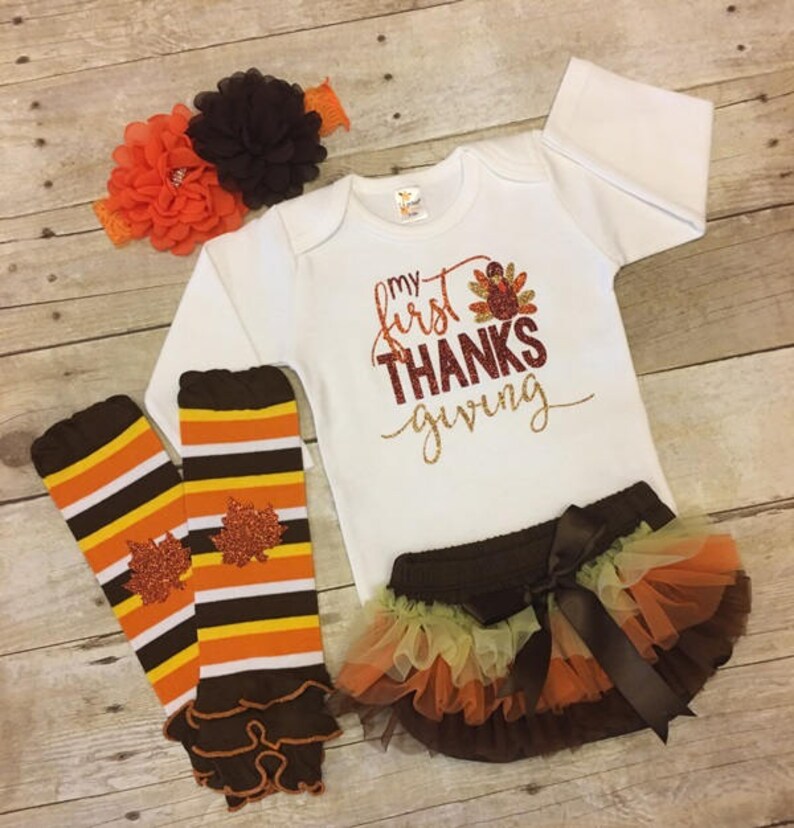 Pu&ograve; includere: Un set di abbigliamento per neonati per il Ringraziamento. Include un body bianco a maniche lunghe con il testo "my first THANKS giving" in glitter dorato, un gambaletto a righe marroni e arancioni con un volant marrone, una gonna di tulle marrone con strati di tulle arancioni e gialli, e una fascia per capelli con fiori marroni e arancioni.
