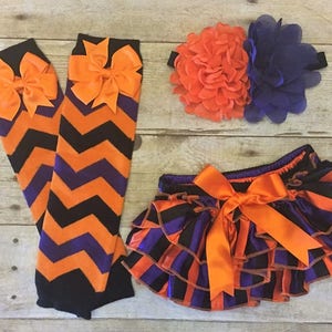 May include: A set of baby girl's clothing in orange, black, and purple. The set includes a headband with a large orange and purple flower, a pair of leg warmers with a chevron pattern, and a pair of bloomers with a striped pattern.
