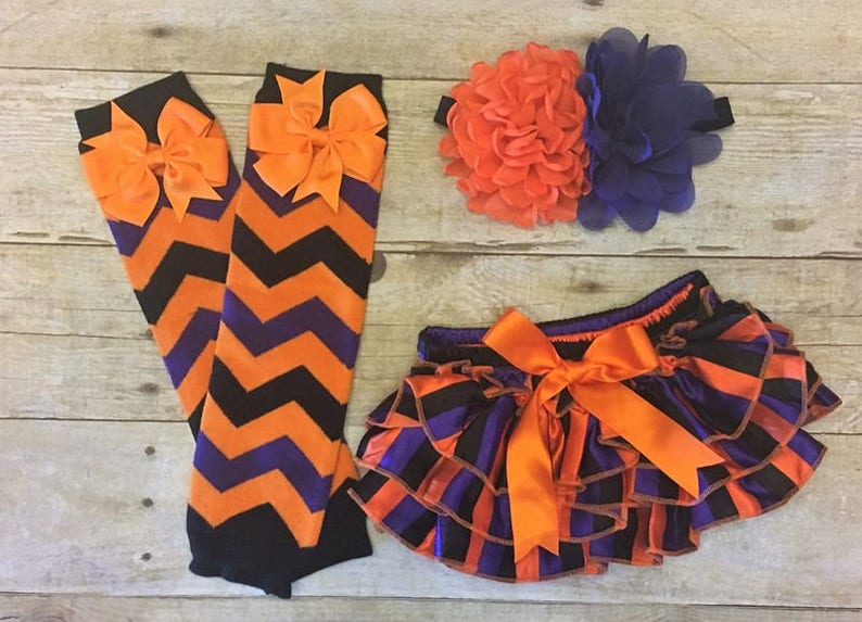 May include: A set of baby girl's clothing in orange, black, and purple. The set includes a headband with a large orange and purple flower, a pair of leg warmers with a chevron pattern, and a pair of bloomers with a striped pattern.