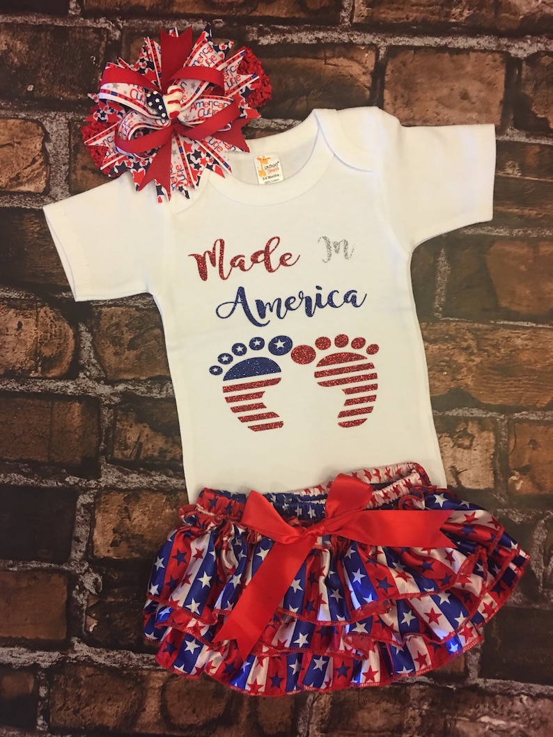 May include: A white baby onesie with the text "Made in America" and red, white, and blue glitter footprints. The onesie is paired with a red, white, and blue star-patterned ruffle skirt with a red bow.