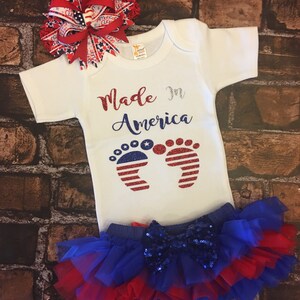 May include: A white baby onesie with the text "Made in America" and a pair of red, white, and blue glitter footprints. The onesie is paired with a blue and red tulle skirt with a blue sequin bow.