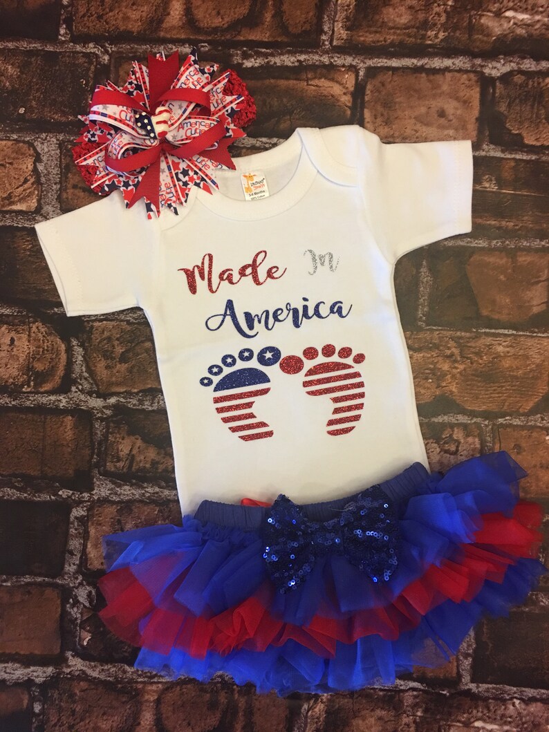 May include: A white baby onesie with the text "Made in America" and a pair of red, white, and blue glitter footprints. The onesie is paired with a blue and red tulle skirt with a blue sequin bow.