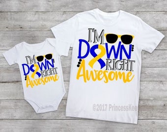 Pode incluir: Duas camisetas brancas com um design de fita azul e amarela. A camiseta maior diz "I'm Down Right Awesome" com óculos de sol. A camiseta menor diz "I'm Down Right Awesome" com óculos de sol.
