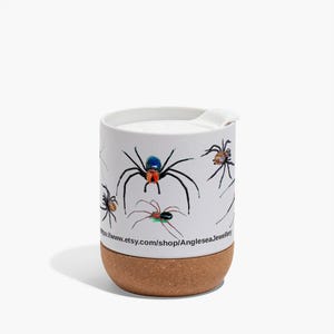 Puede incluir: Taza de viaje de cerámica blanca con fondo de corcho. La taza presenta un diseño colorido de varias arañas sobre un fondo blanco. El texto "https://www.etsy.com/shop/AngleseaJewellery" está impreso en la taza.