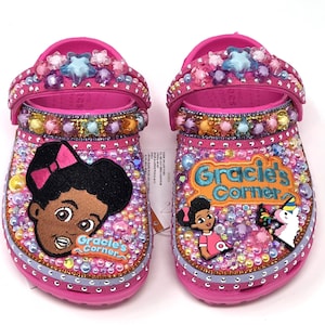 May include: Pair of pink clogs, heavily embellished with colorful rhinestones, beads, and charms. Each clog features a "Gracie's Corner" design, with a cartoon character and text. The straps are adorned with star-shaped and other decorative elements.