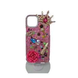 May include: Pink bedazzled phone case with a gold crown, a pink rose, and various decorative gems. The case is covered in small, sparkling crystals and features a lipstick and cocktail glass design.