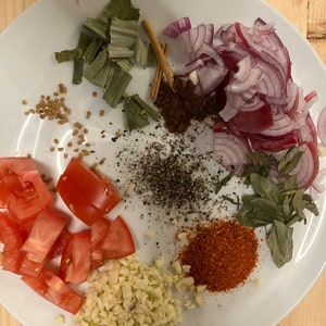 May include: A white plate with various ingredients for cooking, including chopped tomatoes, garlic, red onion, green herbs, spices, and dried herbs.